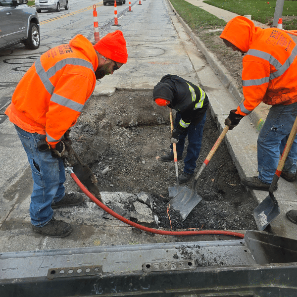 Site Services | Workers in orange safety gear engage in asphalt patching, skillfully using shovels to fill a hole. Traffic cones line the street, and a sidewalk is visible alongside.