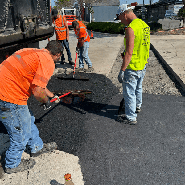 Site Services | Construction workers in orange and yellow vests expertly handle asphalt patching on the road. While one worker operates a tool, others assist to ensure precision. Nearby, a dump truck stands ready in service of the project.