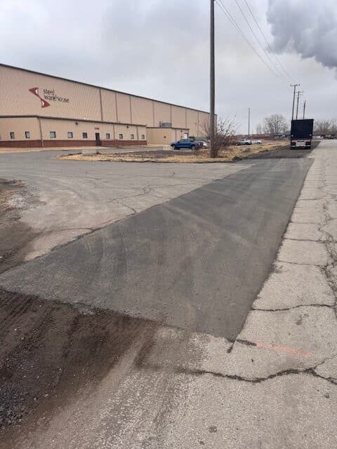 IMG_7691 Site Services | A recently patched section of asphalt road runs alongside a large industrial building with parked cars and a truck under a cloudy sky.