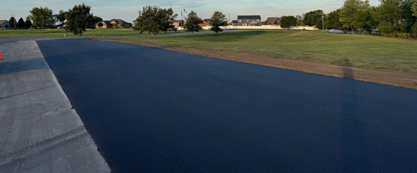 Parking Lot Expansion St John Site Services | A section of freshly paved asphalt paving runs beside a grassy area, with trees and houses in the background under a clear sky.