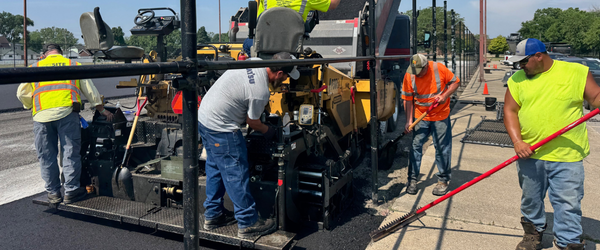 asphalt paving east chicago Site Services | Four construction workers operate paving equipment and smooth fresh asphalt on a road in East Chicago, Indiana, under sunny weather, with traffic cones in the background.